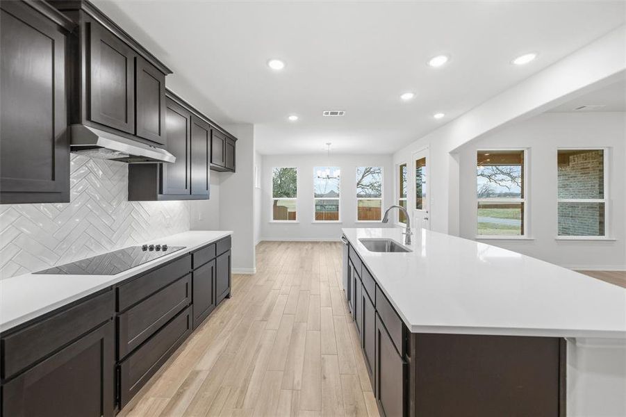 Kitchen with light wood finished floors, backsplash, an island with sink, dark wood finish cabinets, and black electric stovetop