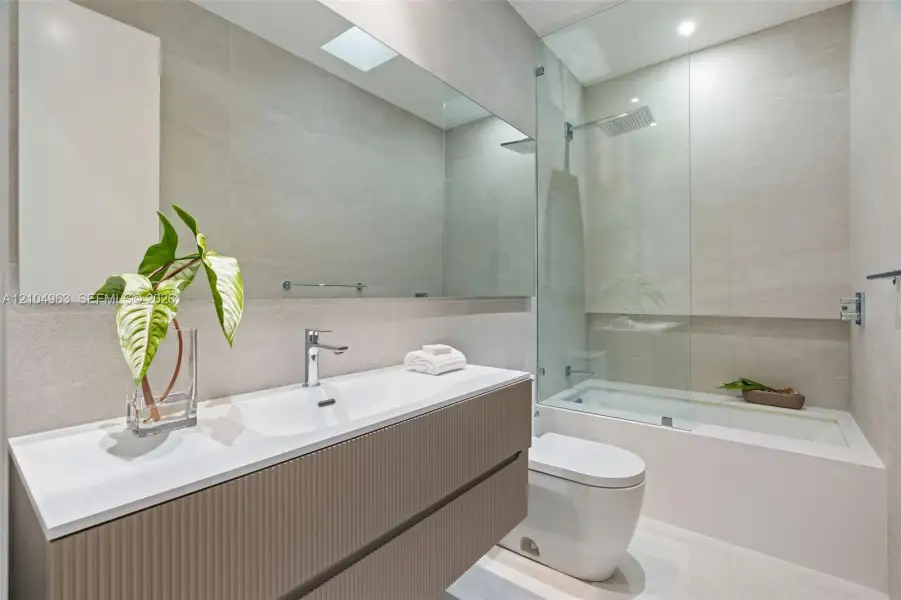 Floating vanity featuring a ribbed textured finish, integrated white basin, wall-to-wall mirror, glass-enclosed shower and tub combination, and full-height neutral wall tile