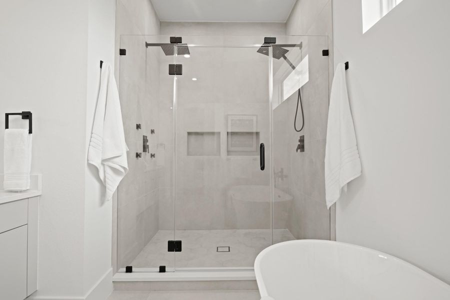 The glass enclosed oversized walk-in shower has hexagonal tile floors and floor-to-ceiling tile walls with dual rain shower heads. One side has a separate spray wand while the other side has four body sprays. Two integrated soap and shampoo shelves complete the look.