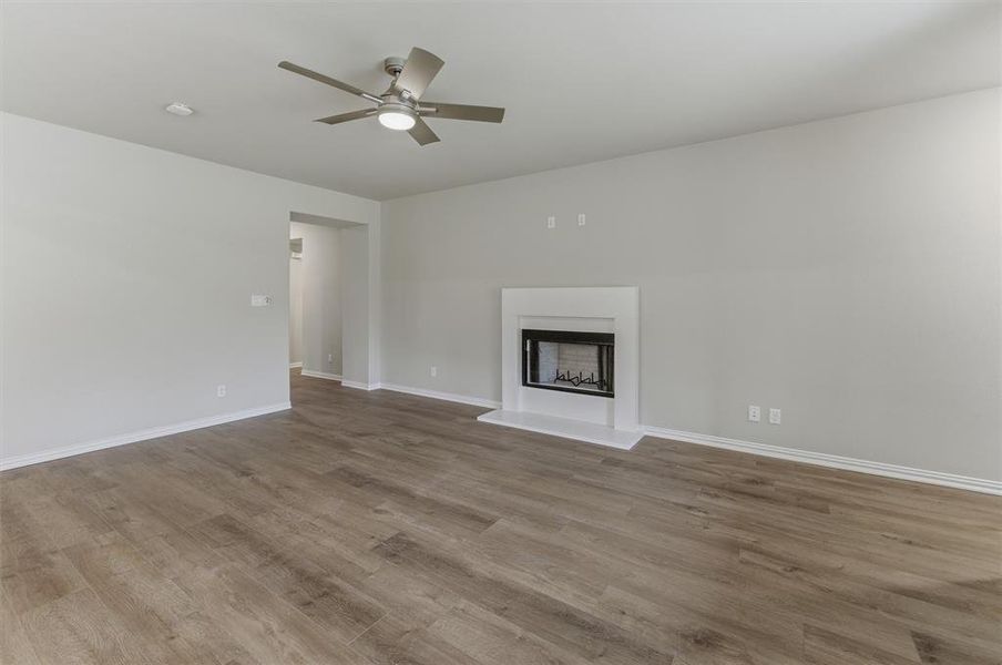 Spacious living area featuring wood-finish flooring, a contemporary ceiling fan with integrated lighting, a built-in fireplace with a white mantel, and light-toned wall finishes