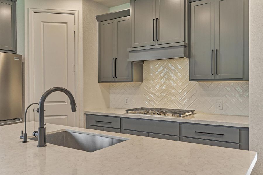 Modern kitchen with sleek gray cabinets, marble countertops, and a stylish herringbone tile backsplash. Features a stainless steel sink with a high-arc faucet and a built-in gas cooktop.