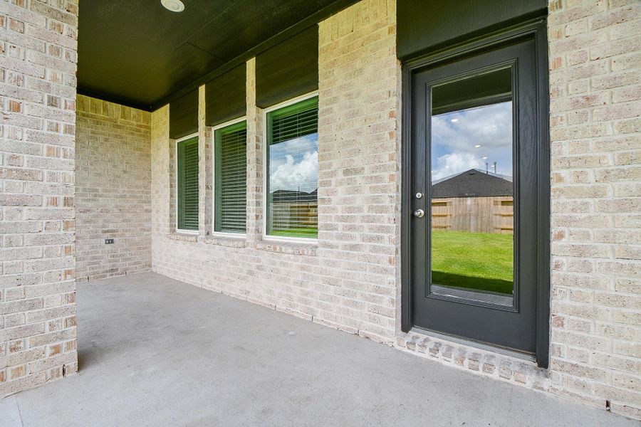 This photo shows a covered patio with light brick walls and a concrete floor. It features a dark-framed glass door and three large windows, offering views of a green backyard with a wooden fence. This photo shows a covered patio with light brick walls and a concrete floor. It features a dark-framed glass door and three large windows, offering views of a green backyard with a wooden fence.