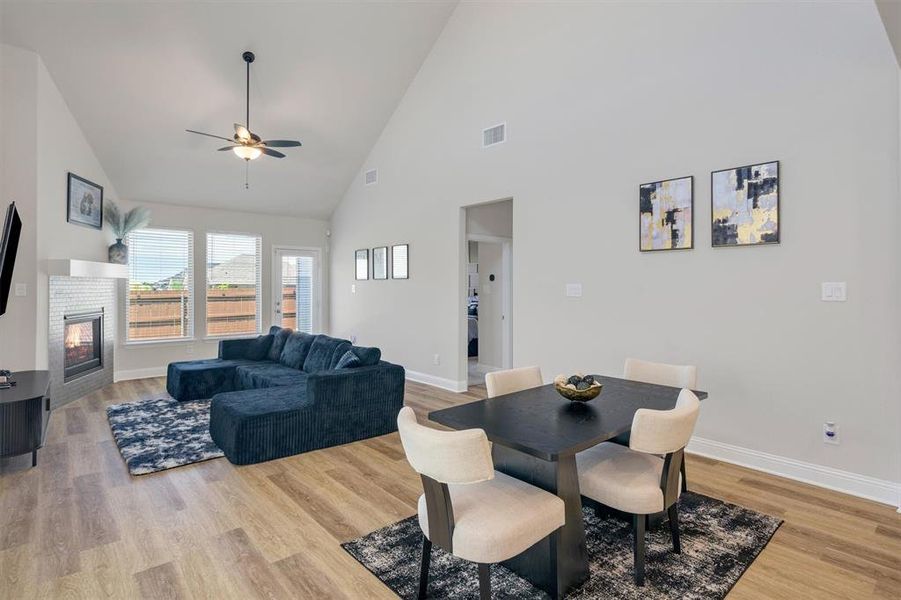 Open-concept living space featuring vaulted ceilings, wood-finish flooring, a fireplace with a decorative mantel and mosaic tile surround, multiple windows, and a glass-paneled door