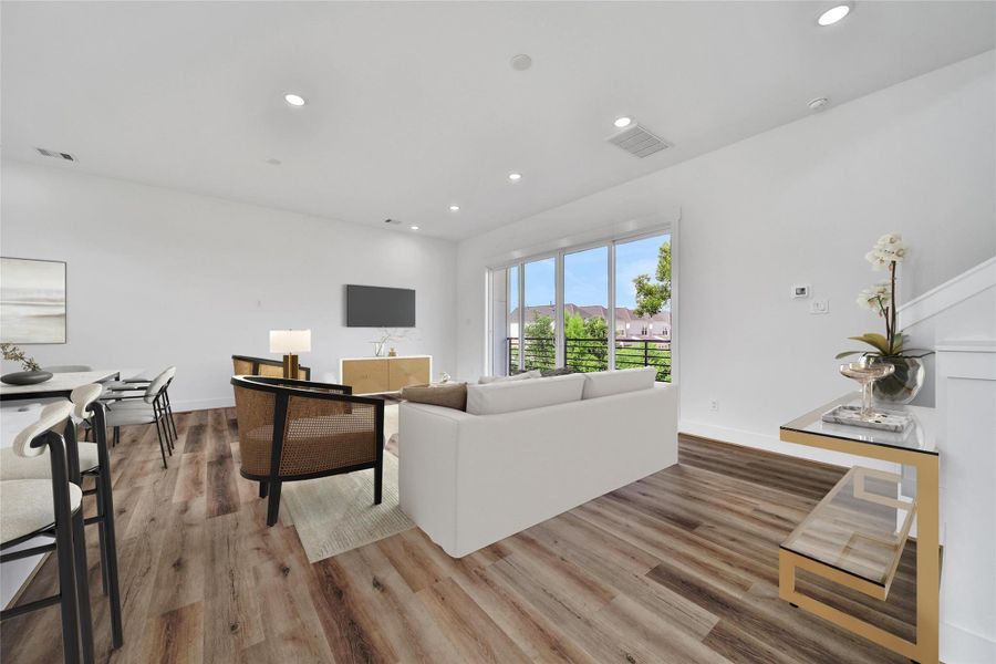 This photo showcases a bright, modern living room with wood flooring and neutral decor. Large windows offer ample natural light and views of the outdoors. The open layout connects to a dining area, creating a spacious, inviting atmosphere.