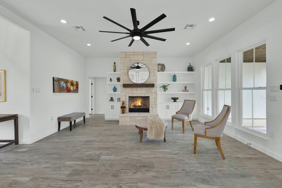 Spacious living area featuring light wood-finish flooring, a stone-clad fireplace with a wood mantle, custom built-in shelving, recessed lighting, and a large ceiling fan with a lot of storage.