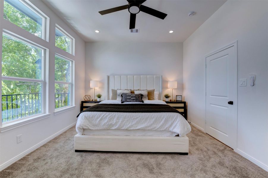 Primary bedroom with vaulted ceiling, recessed lighting, ceiling fan, plush carpet, and oversized windows overlooking mature trees. Balcony access visible on left.