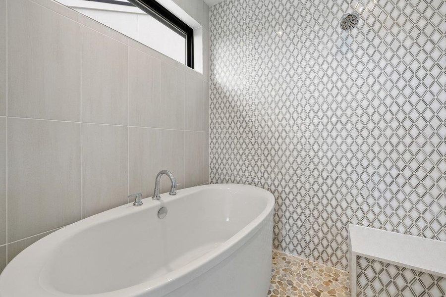 Freestanding soaking tub with chrome fixtures, a geometric mosaic tile shower surround, smooth pebble flooring, and a transom window