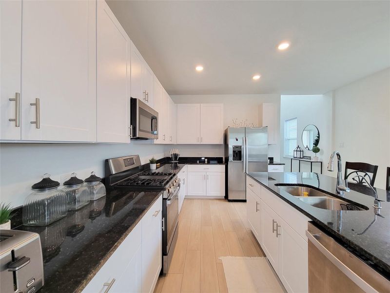 This modern kitchen features sleek white cabinetry, black granite countertops, and stainless steel appliances, including a double oven and fridge. The open layout and ample lighting create a bright, inviting space perfect for cooking and entertaining.