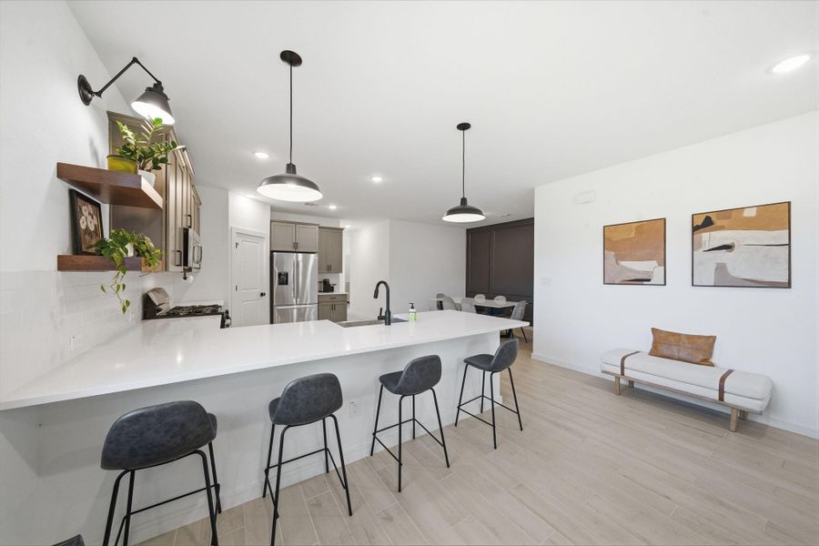 This modern kitchen features a spacious, bright open layout with a large white island and bar seating. It includes stainless steel appliances, stylish pendant lighting, and sleek cabinetry. The adjacent area has contemporary decor, adding to its inviting atmosphere.