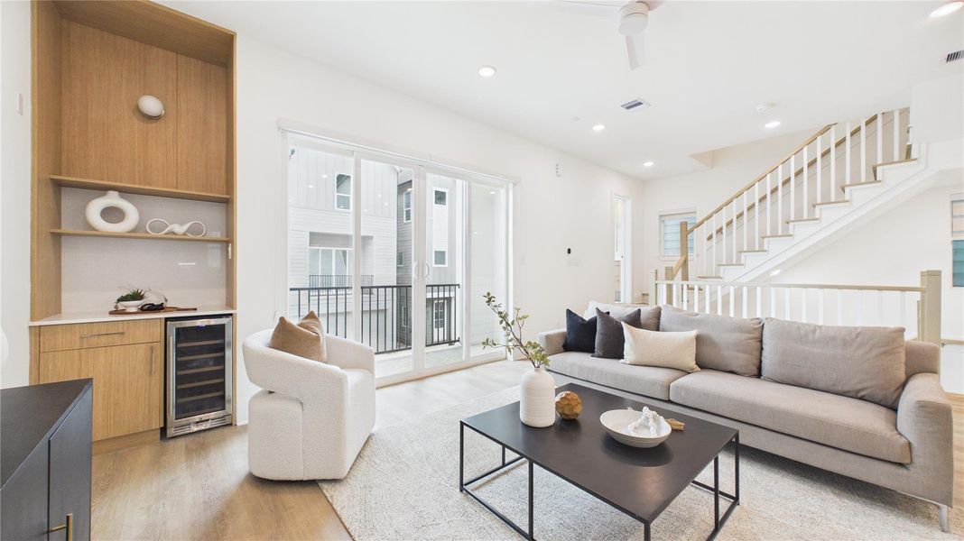 This modern living room features an open layout with natural light streaming through large sliding doors. It includes a cozy gray sofa, a sleek black coffee table, and a built-in wooden shelving unit with a mini-fridge. The space is complemented by light wood flooring and a contemporary staircase.