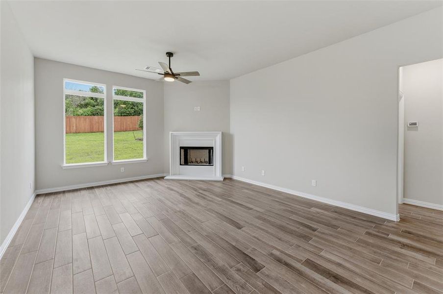Spacious living area featuring wood-finish flooring, a corner fireplace with a white mantel, and two large windows overlooking a green lawn with a privacy fence