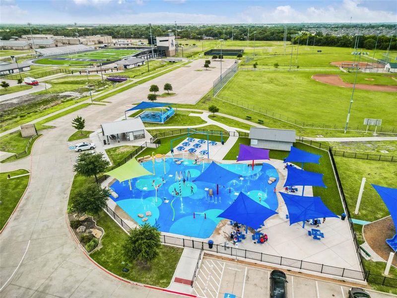 Modern splash pad with multiple water features, shaded seating areas, and a surrounding paved pathway