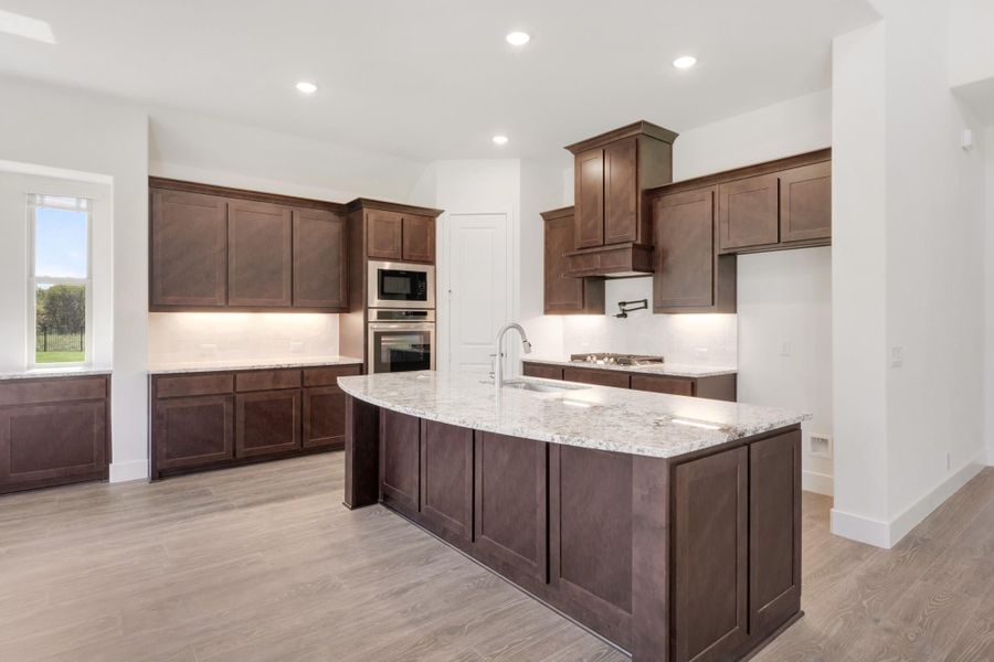 Kitchen with dark wood cabinets, large granite island with sink, and stainless steel appliances on light hardwood floors.