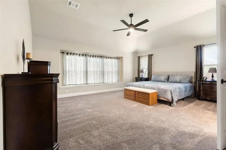 Spacious bedroom featuring vaulted ceilings, neutral-toned carpeting, a contemporary ceiling fan, and multiple windows with blinds Spacious bedroom featuring vaulted ceilings, neutral-toned carpeting, a contemporary ceiling fan, and multiple windows with blinds