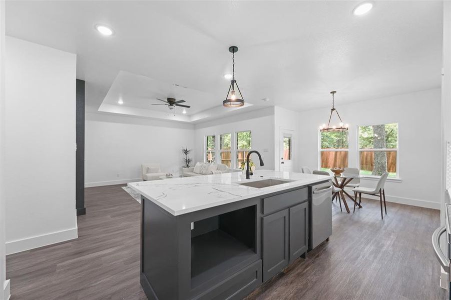 Open-concept living space featuring a central kitchen island with a quartz countertop and integrated sink, pendant lighting, wood-finish flooring, and ample natural light from multiple windows Open-concept living space featuring a central kitchen island with a quartz countertop and integrated sink, pendant lighting, wood-finish flooring, and ample natural light from multiple windows
