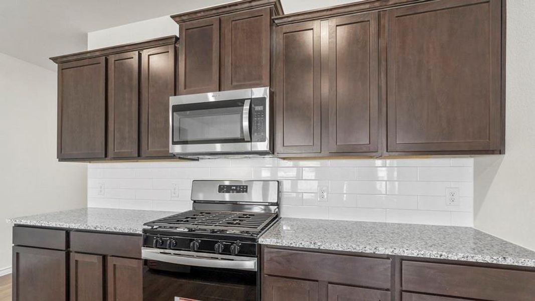 The kitchen features dark wood cabinetry, light speckled countertops, a white subway tile backsplash, and stainless steel appliances, including an oven with a gas range and an overhead microwave