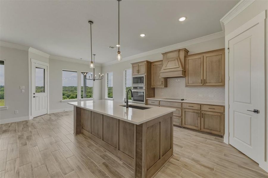 Gourmet kitchen featuring a large island with sink, integrated double ovens, wood-finish cabinetry, light countertops, and wood-finish tile flooring Gourmet kitchen featuring a large island with sink, integrated double ovens, wood-finish cabinetry, light countertops, and wood-finish tile flooring