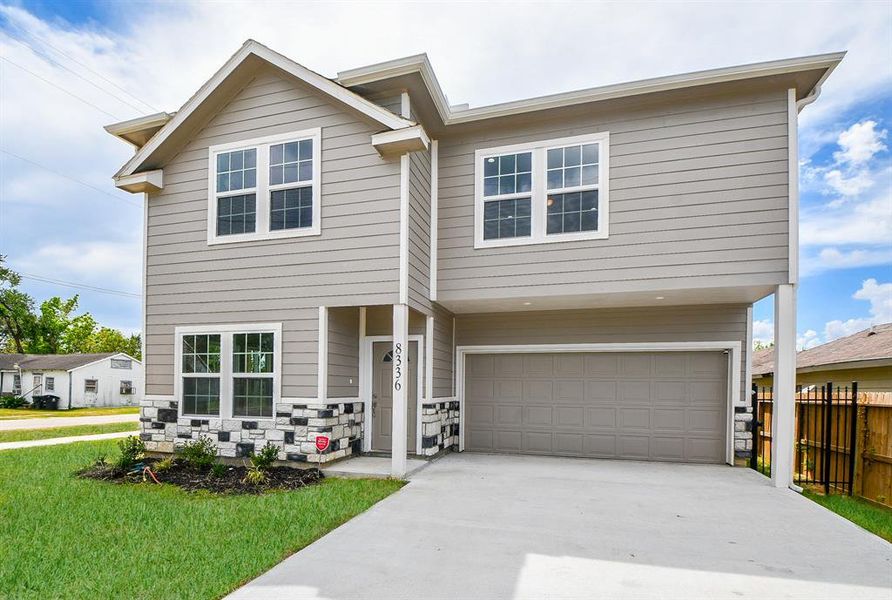 This is a modern two-story  corner home featuring a neutral gray siding with stone accents around the garage and entryway. It includes a two-car garage and has a freshly landscaped yard under a bright, cloud-dotted sky.