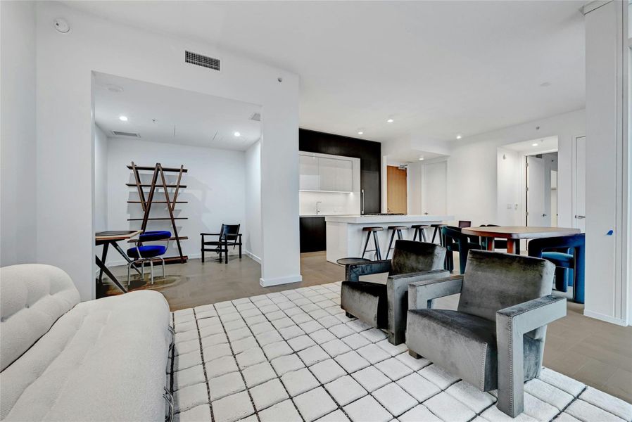 This modern living area features an open floor plan that seamlessly connects to a versatile DEN, ideal for a home office, reading nook, or additional seating area. The room is bright and airy, with clean lines, neutral tones, and contemporary furnishings that create a welcoming and stylish atmosphere. The nearby kitchen is visible from the living area, offering a cohesive and functional layout perfect for both everyday living and entertaining. Furnishings provided by Scott + Cooner available for purchase separately. Photo is model unit #2406.