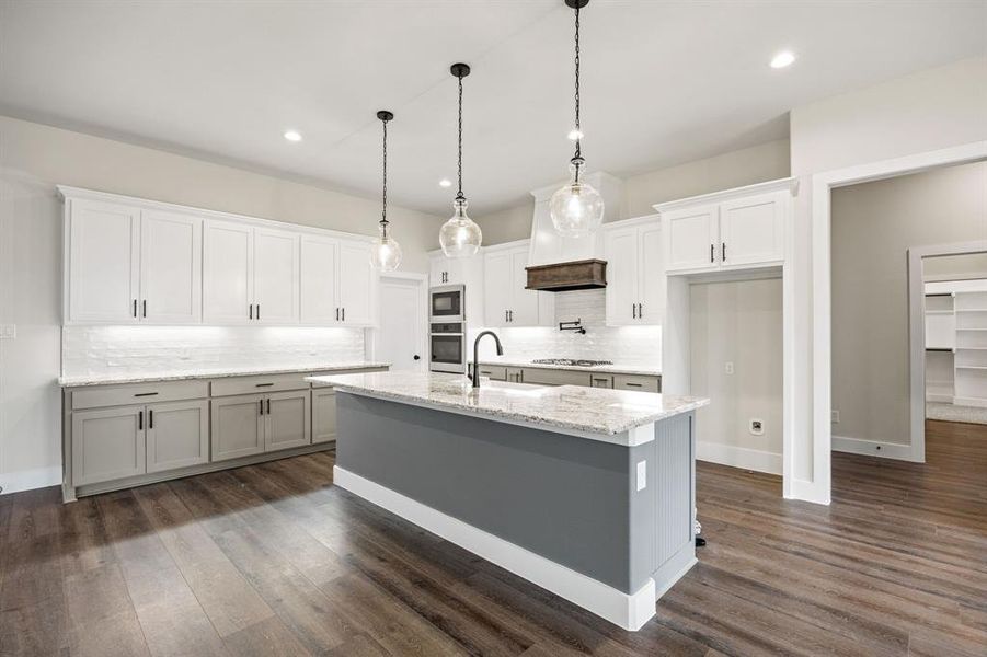 Two tone kitchen featuring light stone countertops, pendant lighting, backsplash, a kitchen island with sink, and dark wood-style floors Two tone kitchen featuring light stone countertops, pendant lighting, backsplash, a kitchen island with sink, and dark wood-style floors