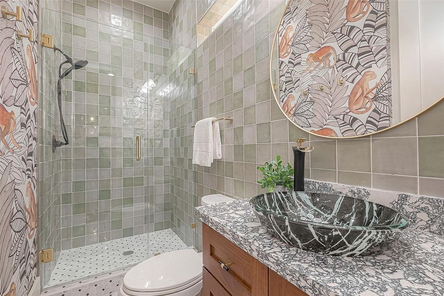 This photo showcases a modern bathroom with a glass-enclosed shower, a distinctive vessel sink atop a granite vanity, and a whimsical wallpaper accent wall. The space is complemented by gold-toned fixtures and a round mirror with a matching gold trim. This photo showcases a modern bathroom with a glass-enclosed shower, a distinctive vessel sink atop a granite vanity, and a whimsical wallpaper accent wall. The space is complemented by gold-toned fixtures and a round mirror with a matching gold trim.