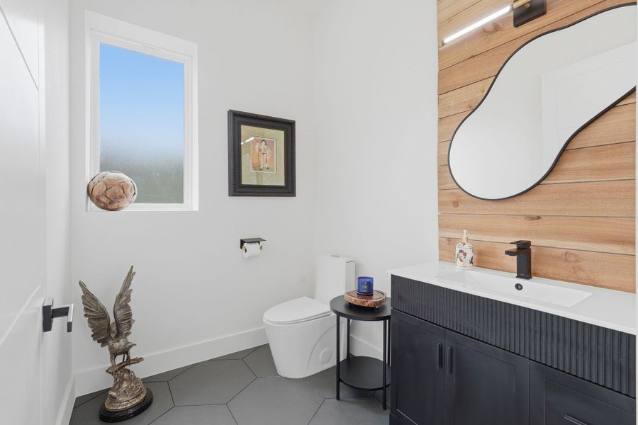 This bathroom features a modern design with a unique mirror, wood accent wall, and hexagonal floor tiles. It includes a sleek black vanity, a window for natural light, and stylish decor elements.