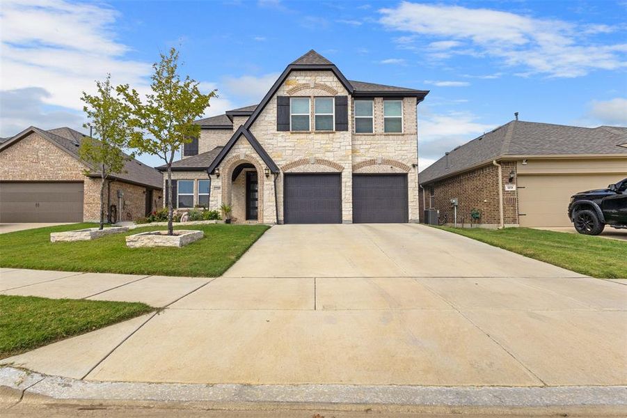 French country style house featuring stone siding, concrete driveway, a front lawn, roof with shingles, and an attached garage
