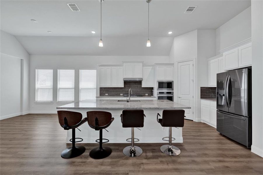 Kitchen featuring stainless steel appliances, white cabinets, a kitchen island with sink, light stone counters, and lofted ceiling