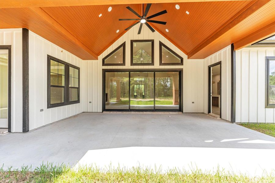 This photo showcases a spacious, covered outdoor patio with a ceiling fan and wood-paneled ceiling. Large sliding glass doors connect the space to the interior, while an adjacent area features an outdoor kitchen, ideal for entertaining. The patio overlooks a lush, green lawn surrounded by trees.