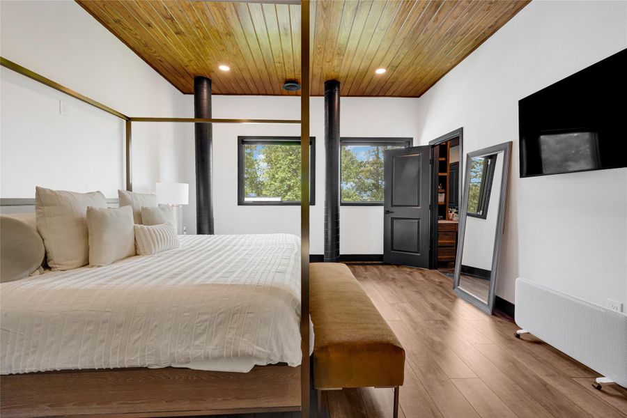 This bedroom features a modern design with a canopy bed, wood-paneled ceiling, and large windows providing natural light. The room includes a wall-mounted TV, stylish black accents, and an open doorway leading to additional space, creating a warm and inviting atmosphere.
