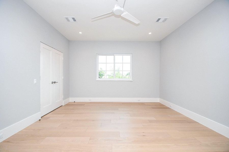 A bright, minimalist room with light wood flooring and soft gray walls. It features a window bringing in natural light, a ceiling fan, and a double-door closet, creating a clean and modern feel.