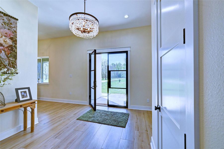 Bright entryway featuring a modern glass door with a view of lush greenery. The space includes light wood flooring, a decorative chandelier, and a welcoming side table.