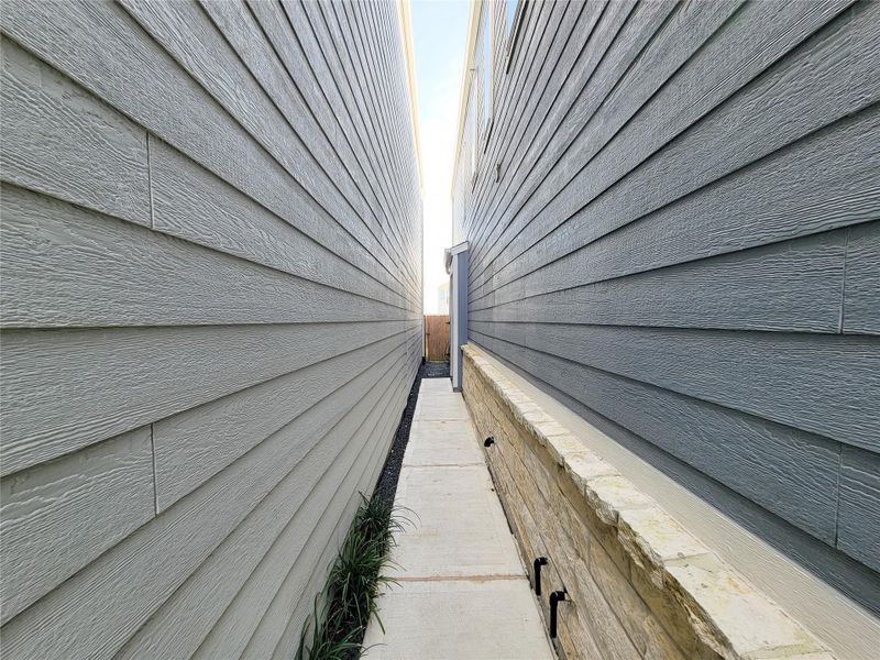 Narrow alleyway between two houses with wood siding. The pathway is concrete, bordered by a low stone wall on the right, leading to a fenced area in the distance.