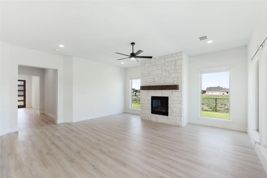 Open living room with stone fireplace, wood mantel, ceiling fan, and light hardwood floors