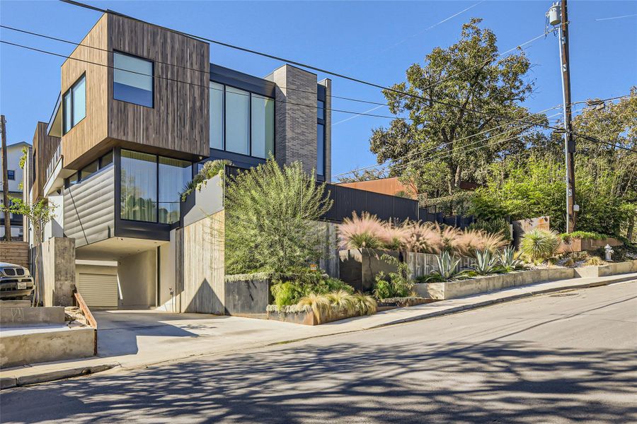 Modern architecture with wood and concrete accents features lush, multi-level landscaping along a sloped street. Modern architecture with wood and concrete accents features lush, multi-level landscaping along a sloped street.