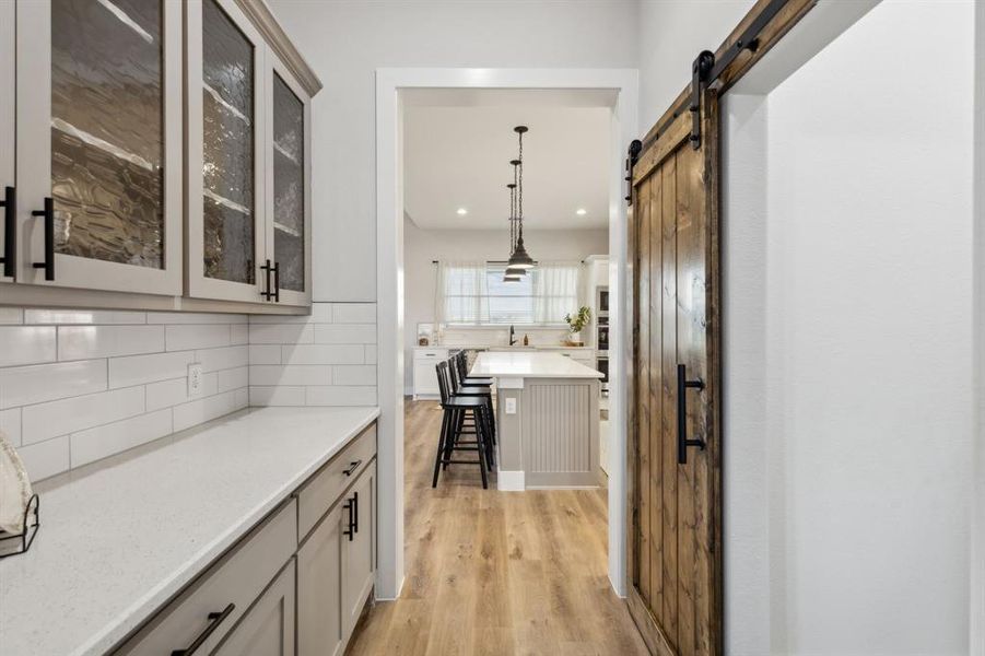 Modern kitchen pantry featuring a wood-finish barn door with black hardware, light-toned cabinetry with glass inserts, a white subway tile backsplash, and light-toned countertops