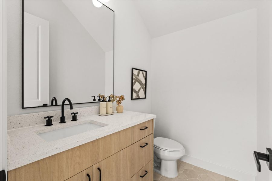 The main-floor powder room features a warm wood vanity with a white quartz countertop, an under-mount sink, and matte black hardware. A black gooseneck faucet pairs with a matching black-framed mirror, set against neutral vertical walls and geometric floor tiles.