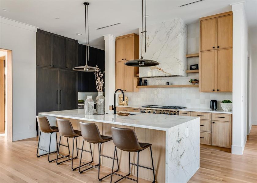 Kitchen featuring light hardwood / wood-style flooring, a kitchen island with sink, tasteful backsplash, light stone countertops, and decorative light fixtures