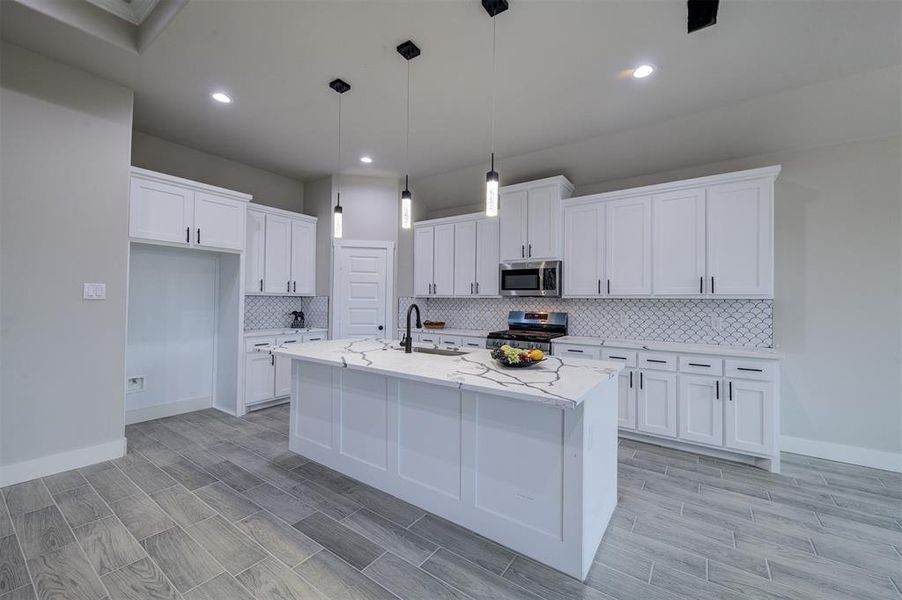 Kitchen with decorative light fixtures, an island with sink, stainless steel appliances, light stone countertops, and white cabinets