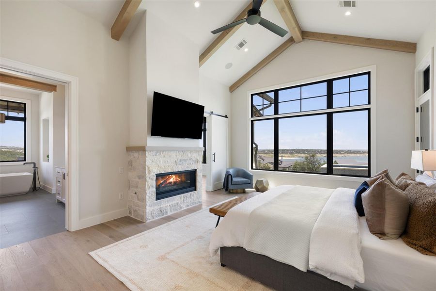 Bedroom featuring beamed ceiling, high vaulted ceiling, a glass covered fireplace, light wood-type flooring, and a ceiling fan