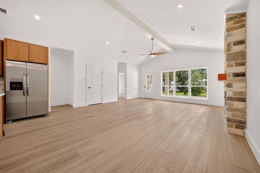 Open-concept living space featuring vaulted ceilings with exposed beams, wood-finish flooring, a stone-clad fireplace, and large windows