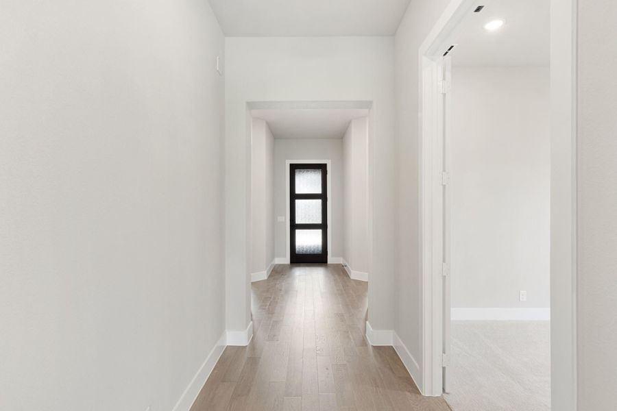 Hallway with light hardwood floors, white walls, and black glass-panel door at the end