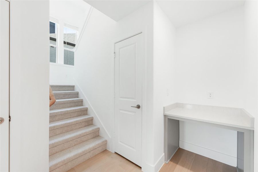 Hallway with carpeted staircase, white walls, and built-in desk on light hardwood floors