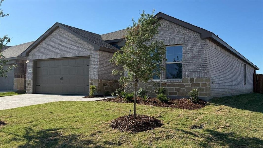 Single-story exterior featuring a light gray brick facade with stone accents, an attached garage with a taupe door, a concrete driveway, and a front yard with a young tree and landscaping