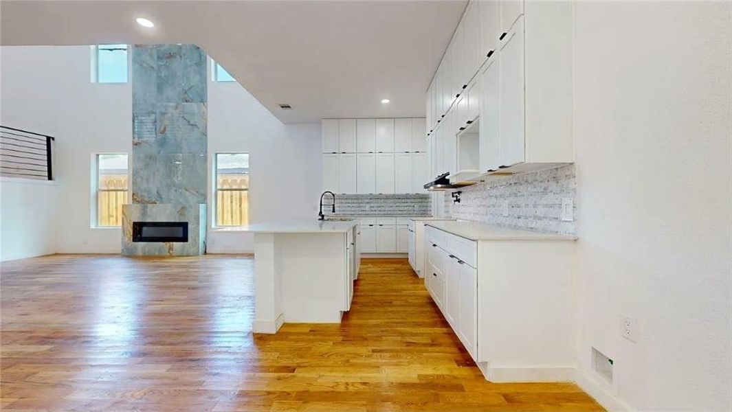 Open-concept kitchen and living area featuring wood-finish flooring, a contemporary fireplace with a stone surround, white cabinetry, a kitchen island, and a mosaic tile backsplash