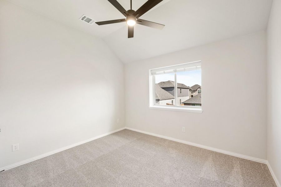 Empty bedroom with vaulted ceiling, ceiling fan, carpet flooring, and a window overlooking neighboring homes