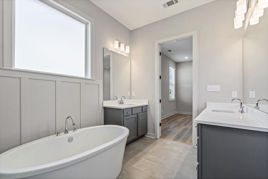 Freestanding soaking tub with chrome fixtures, dual vanities with white countertops, board and batten wainscoting, and large tiled flooring