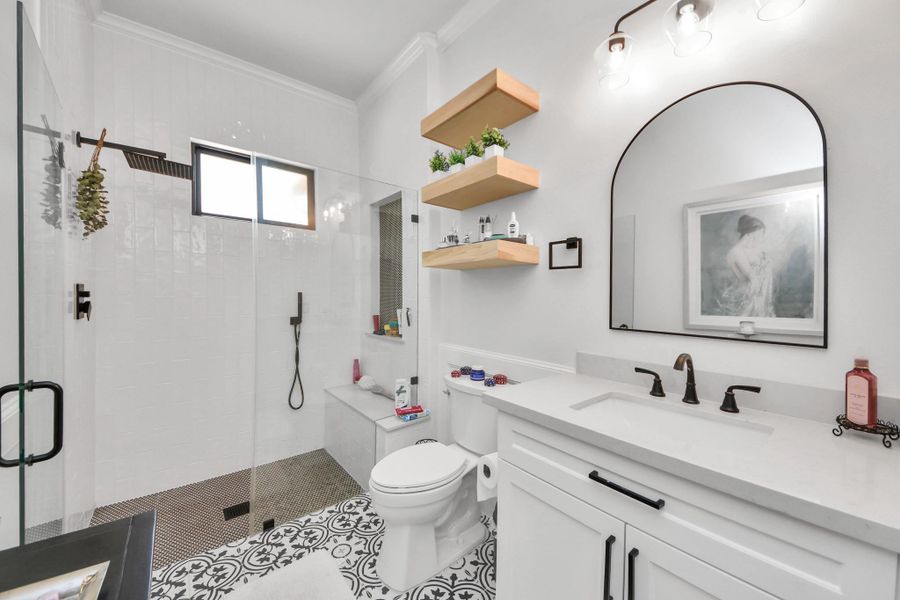 This bathroom features a modern design with a glass-enclosed shower, patterned tile flooring, and a sleek vanity with a black-framed mirror. Open shelving adds storage and a touch of greenery, while sophisticated lighting enhances the space.