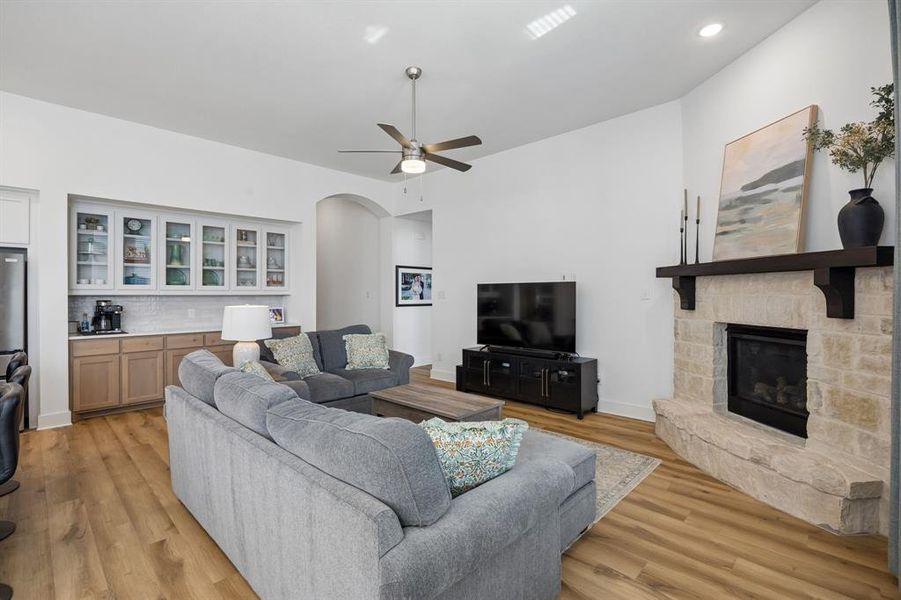 Open-concept living area featuring wood-finish flooring, a stone fireplace with a dark wood mantel, high ceilings, and a built-in wet bar with glass-front cabinetry and a tile backsplash