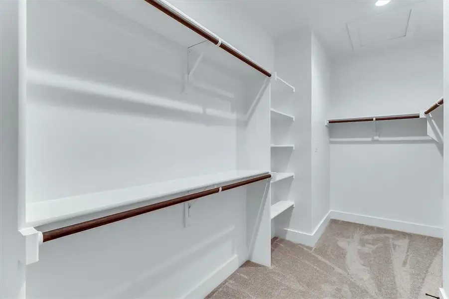 Spacious walk-in closet featuring white built-in shelving, dark wood-finish hanging rods, and light-colored carpet flooring Spacious walk-in closet featuring white built-in shelving, dark wood-finish hanging rods, and light-colored carpet flooring